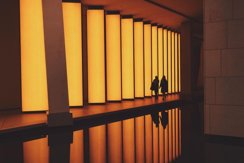 Free Image of Couple Walking Down Hallway Next to Tall Windows 