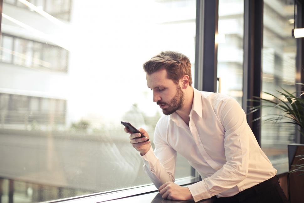 Free Image of Young Adult Man in White Shirt Holding Phone Near Window 