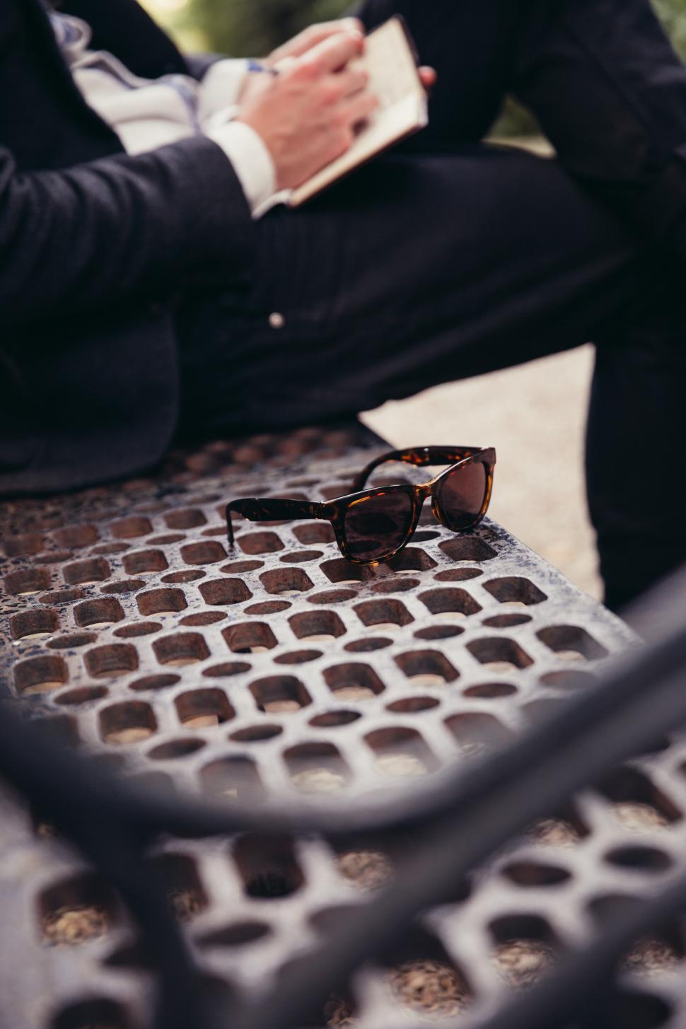 Free Image of A caucasian man writing and sunglasses kept on a park bench 