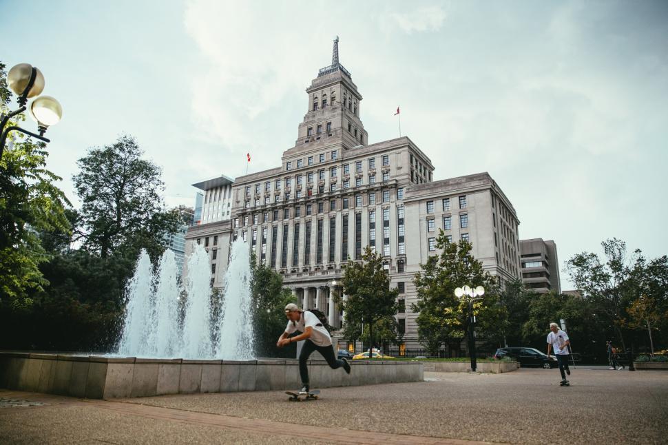 Free Image of Young Caucasian skateboarders ride past a fountain 