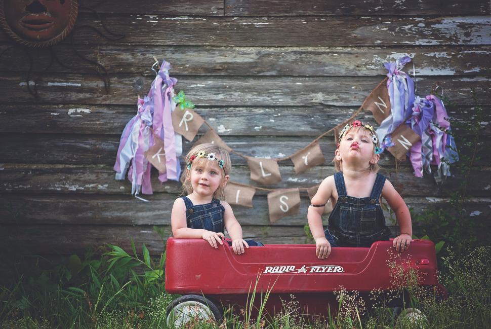 Free Image of Cute Little Twin Sisters Sitting in Wagon Toy Against Wooden Plank Wall 