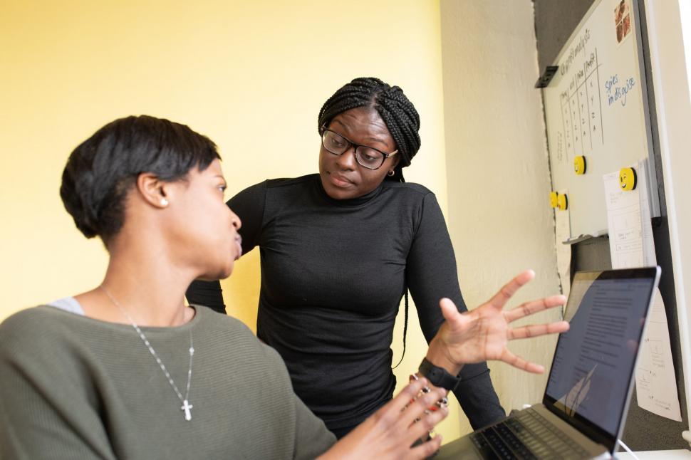 Free Image of Two Young Women Discussing at office desk with whiteboard and laptop 