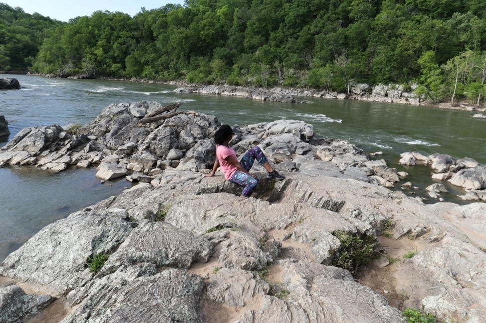 Free Image of Young Woman in yoga pants sitting on rock with river and trees in the background 