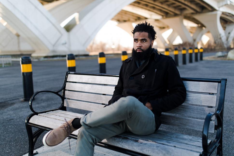 Free Image of Young Man in black jacket on bench with road and bridge - looking at away 