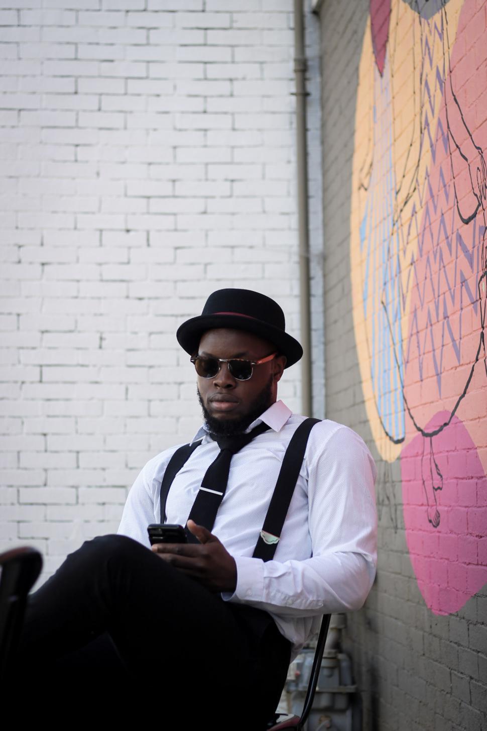 Free Image of Young Man wearing suspenders and hat using cell phone against graffiti wall 