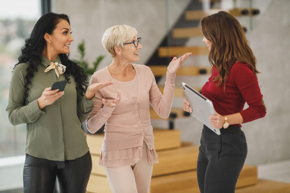 Free Image of Three female coworkers talking and smiling while standing in the middle of a workspace 