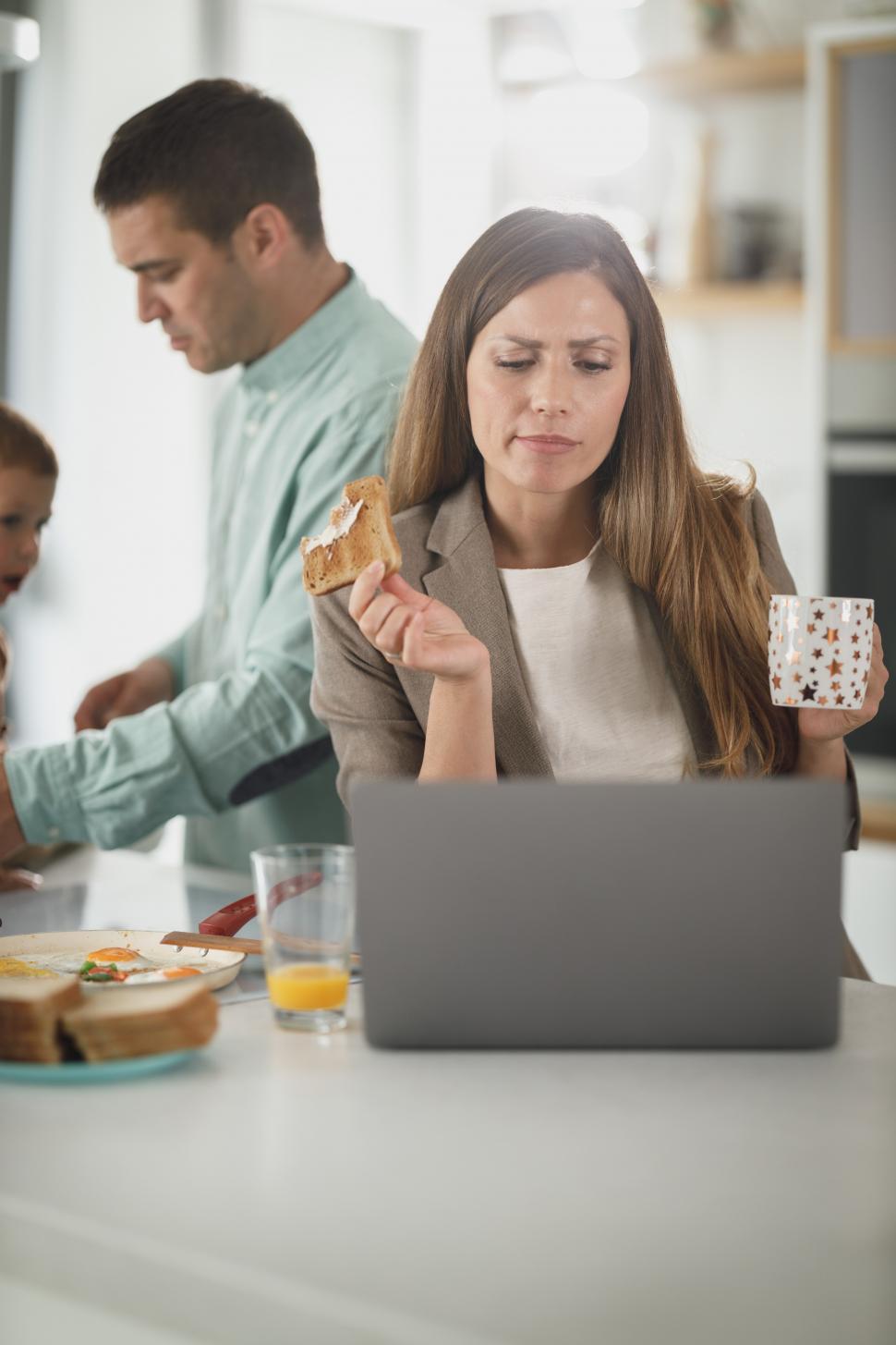 Free Image of Young mother holding coffee cup and eating toast while using laptop in the kitchen 
