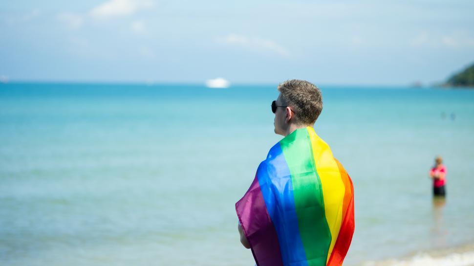 Free Image of LGBT travel concept. Man on the beach draped in Pride flag 