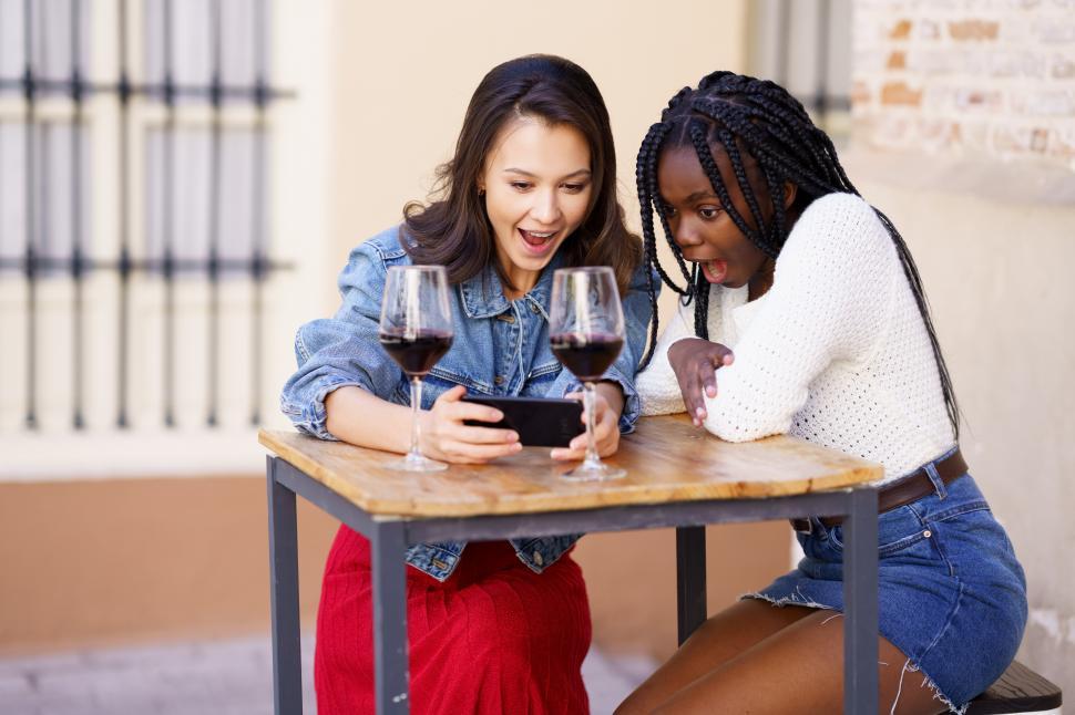 Free Image of Two women looking at their smartphone together while having a glass of wine. 