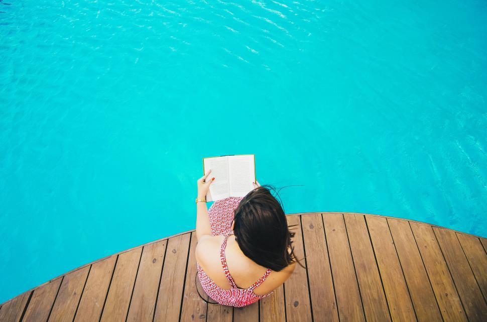 Free Image of A woman sitting on a deck by a pool reading a book 