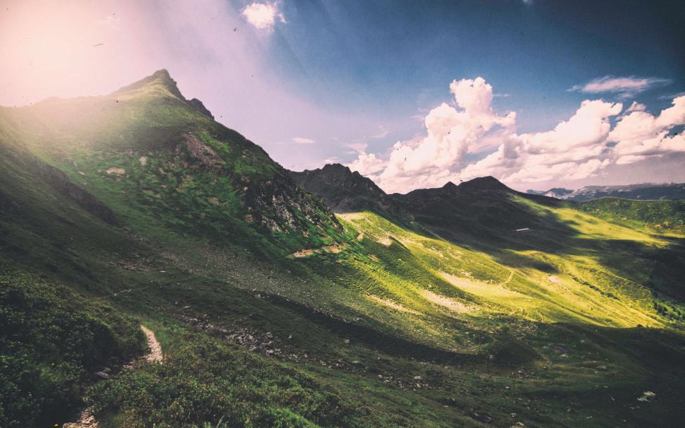 Free Image of A green hills with a path and clouds in the sky with quiraing in the background 