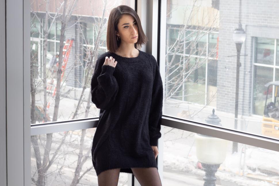 Free Image of Woman in black sweater standing by a large window, indoors 