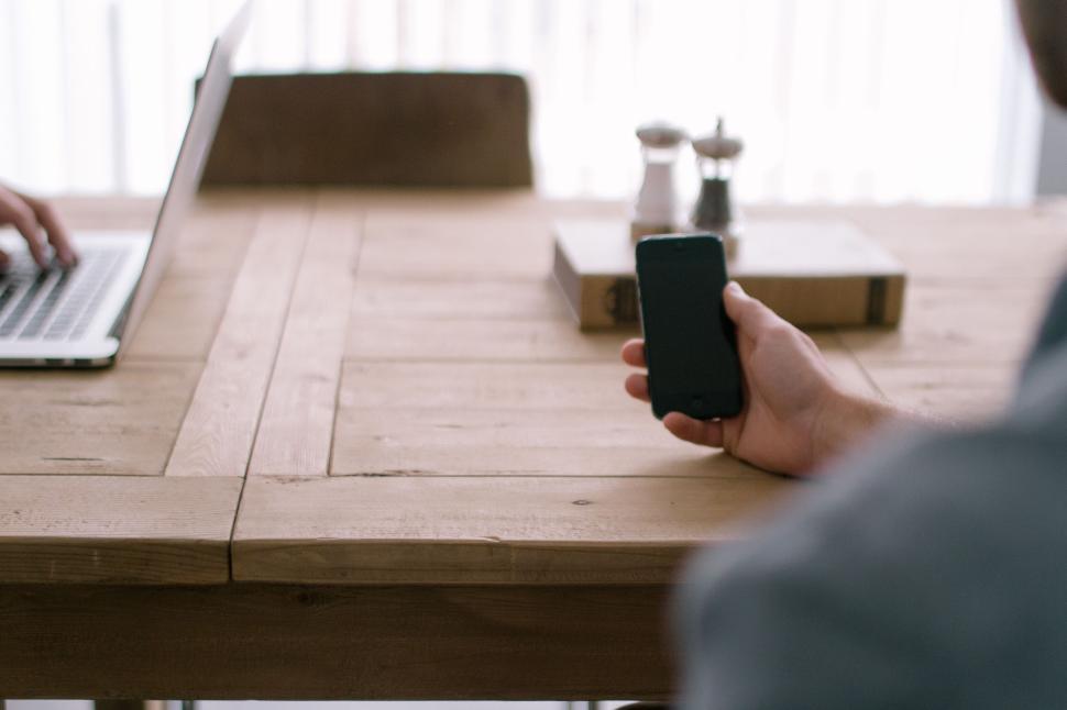 Free Image of Man holding smartphone next to laptop on wooden table. 