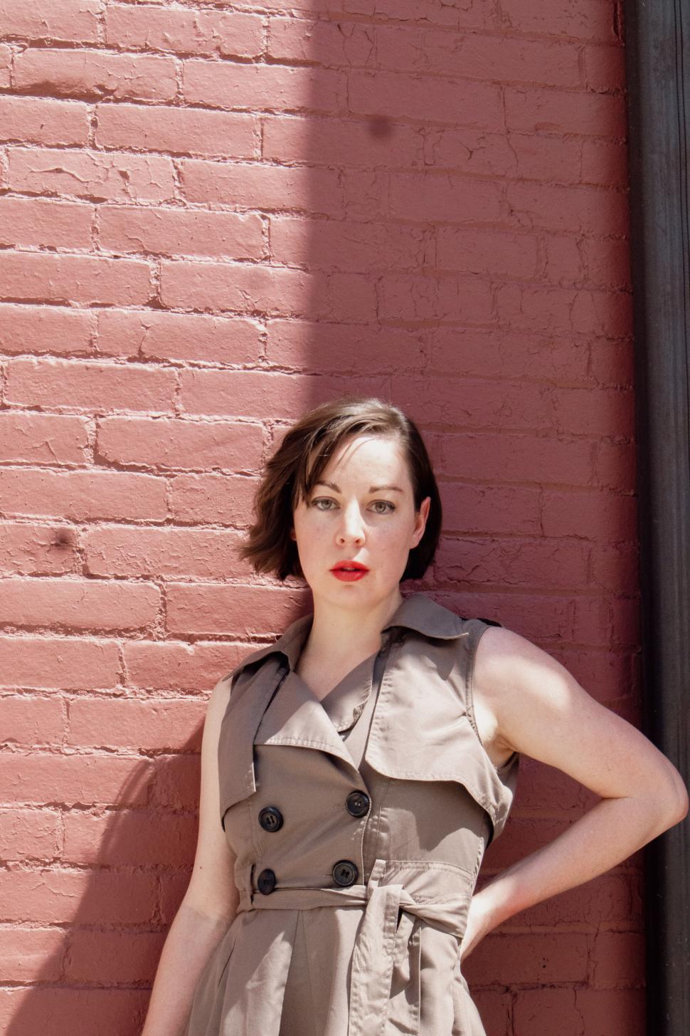 Free Image of Woman in beige trench coat standing against red brick wall 
