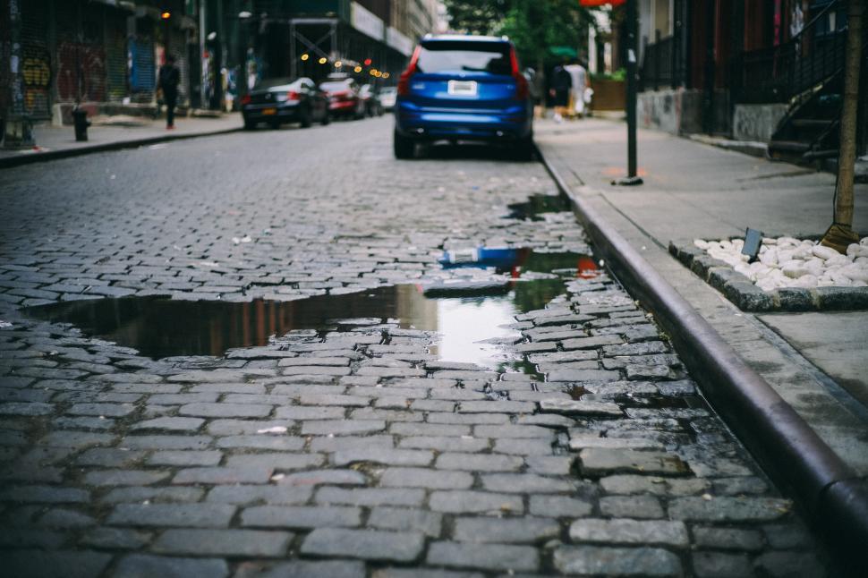 Free Image of Wet cobblestone street with puddle and parked vehicle. 