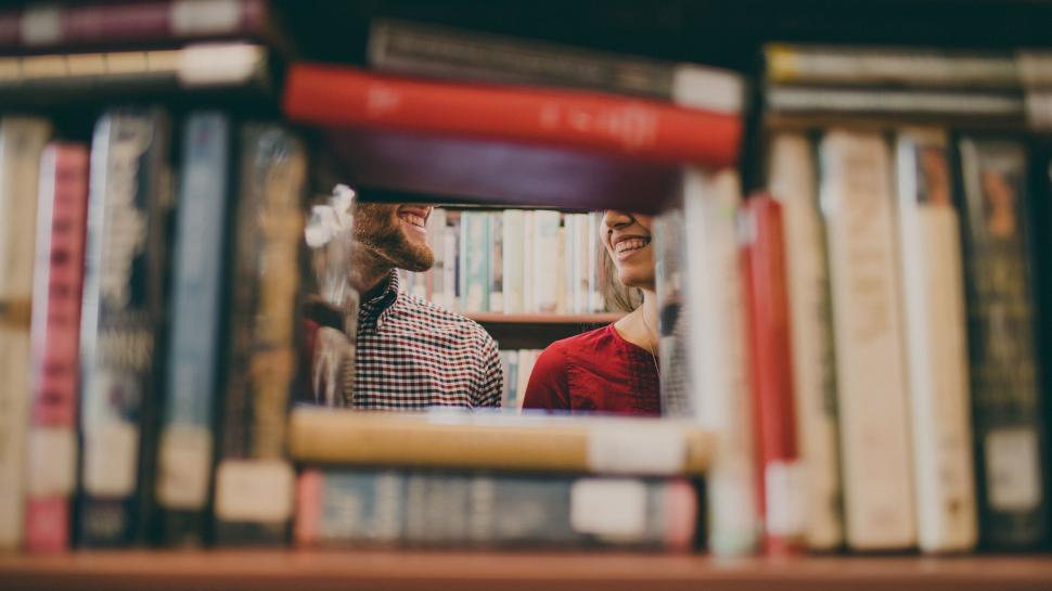 Free Image of Two people peeking through a bookshelf in a library. 