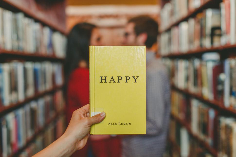Free Image of Person holding a book titled Happy in a library aisle. 