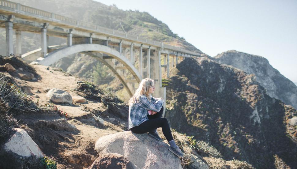 Free Image of Person sitting on a rock near a large bridge structure. 