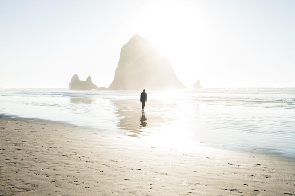 Free Image of Solitary figure walks along a serene beach with sun lit rock in background. 
