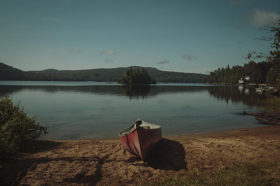 Free Image of Serene lakeside scene with a red canoe on a sandy shore. 