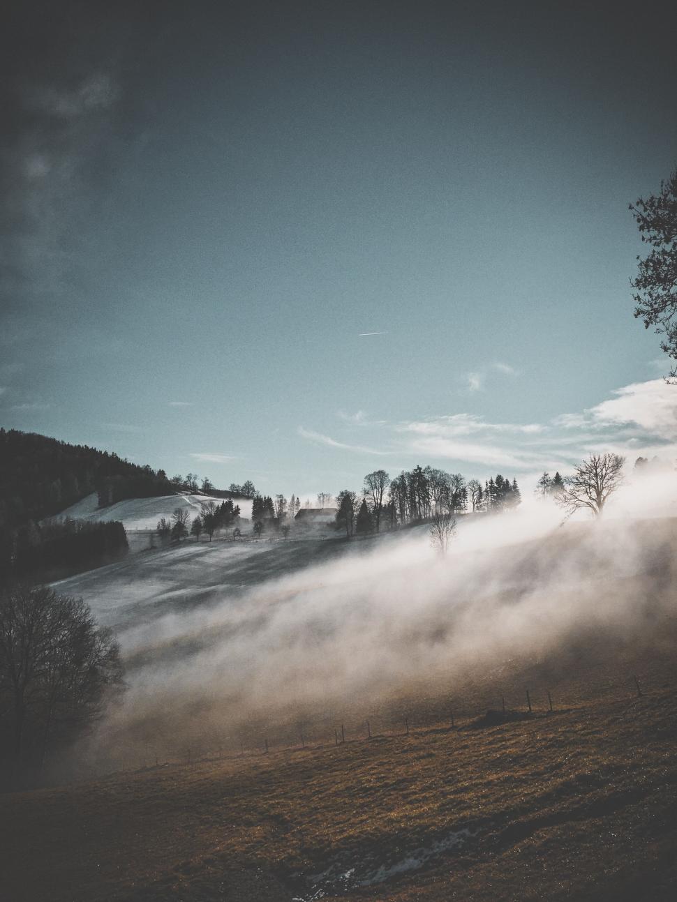 Free Image of Misty morning hillside with trees and fog under blue sky 