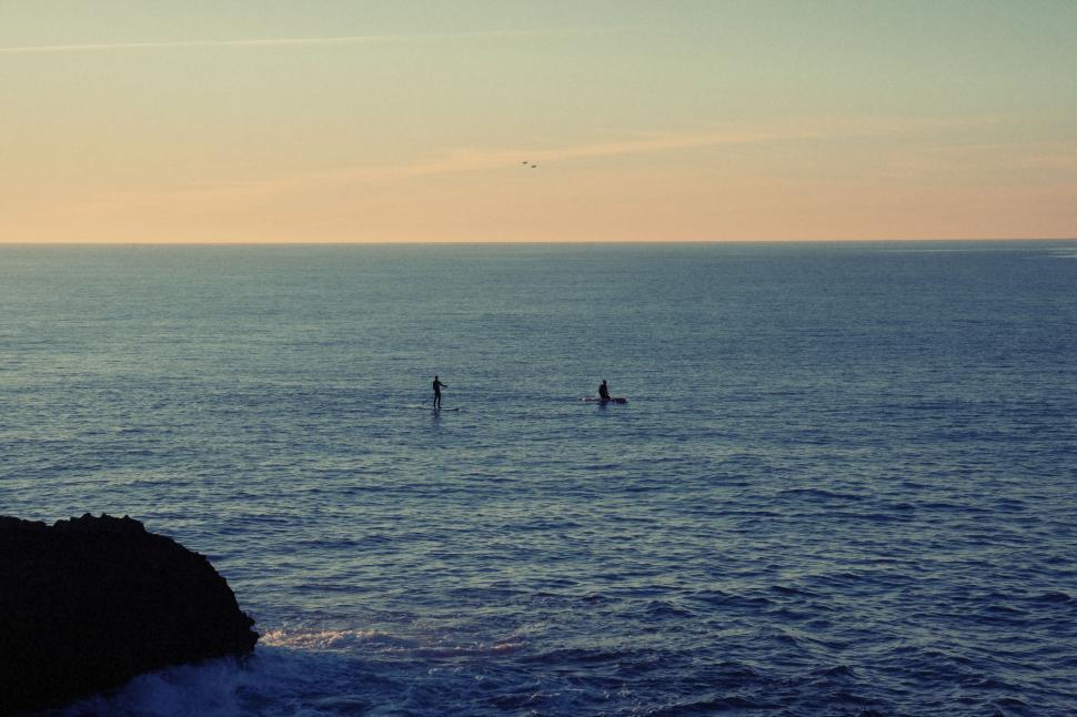 Free Image of Sunset over a calm ocean with two silhouetted paddle boarders visible 
