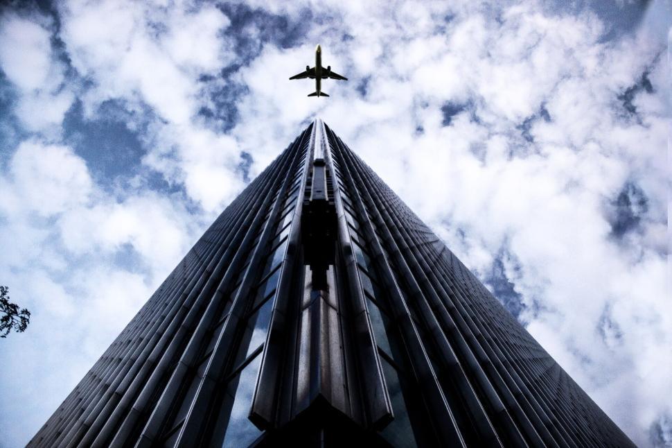 Free Image of Aeroplane flying over a modern skyscraper with cloudy background. 