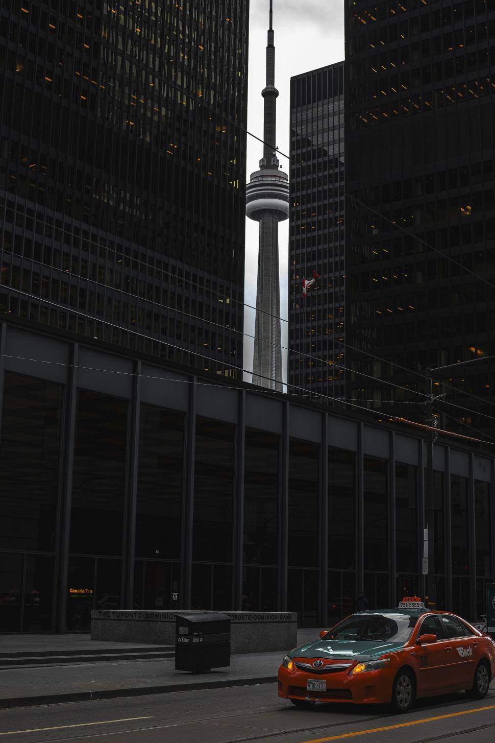 Free Image of CN Tower framed by tall buildings with an orange taxi in front. 