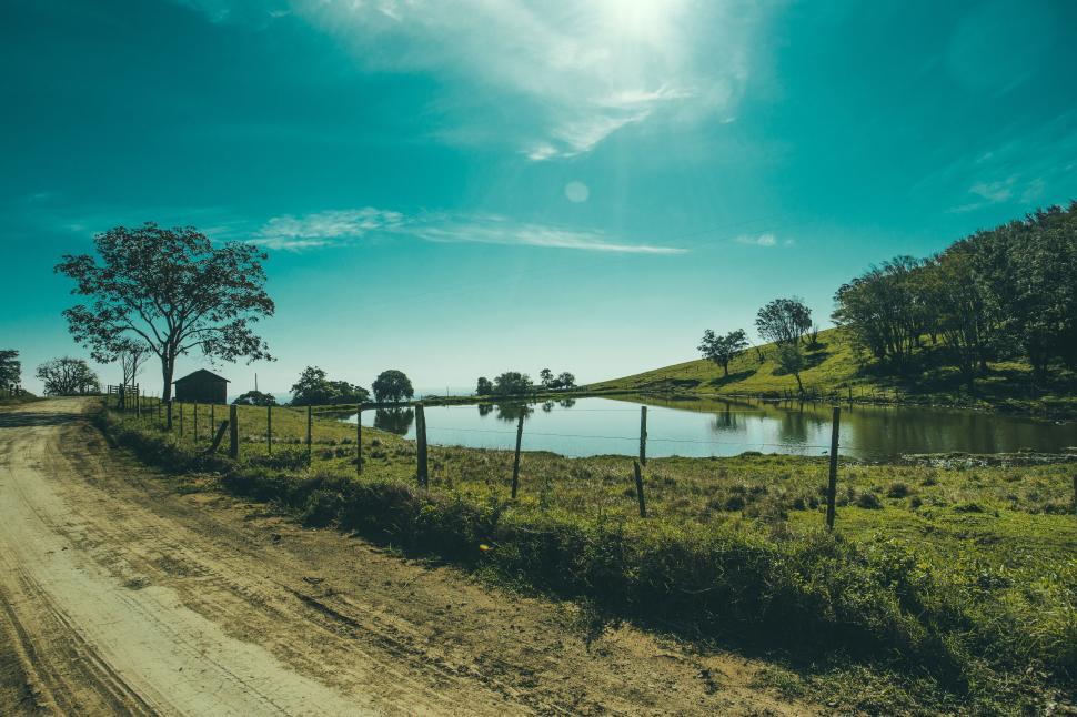Free Image of Dirt road by the lake under blue sky with trees and fencing. 