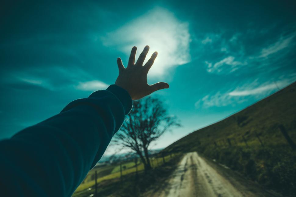 Free Image of Person reaching towards bright sun on rural path with tree ahead. 