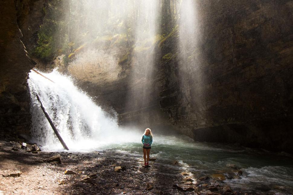 Free Image of Person standing near waterfall with sun rays filtering through trees. 