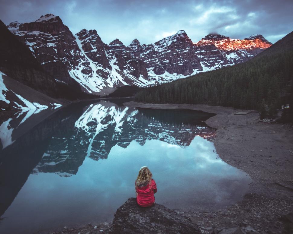 Free Image of Person in red jacket sits by tranquil lake with snow-capped mountains. 