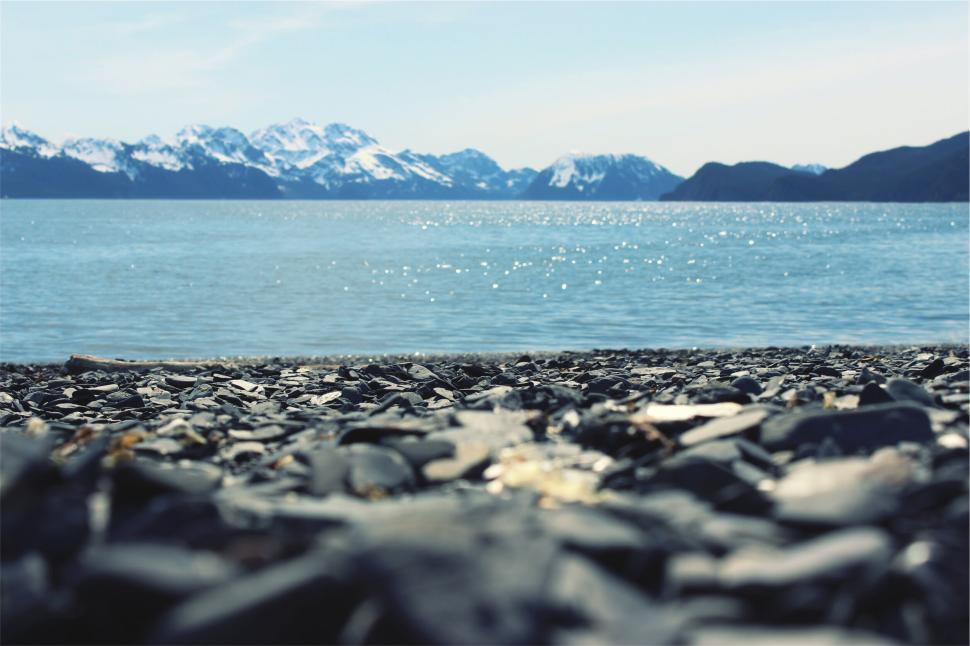 Free Image of Rocky beach under a bright sky with snowy mountains in the distance. 