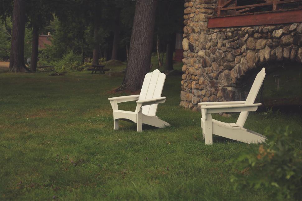 Free Image of Two white Adirondack chairs on green grass near a rustic stone house 