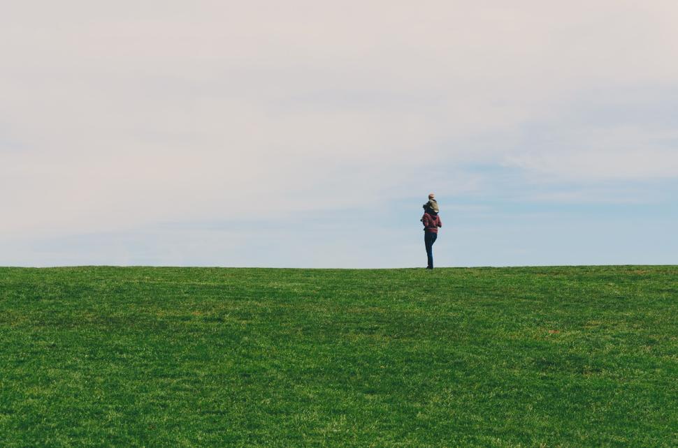 Free Image of Person walking across a vast green field under a blue sky. 
