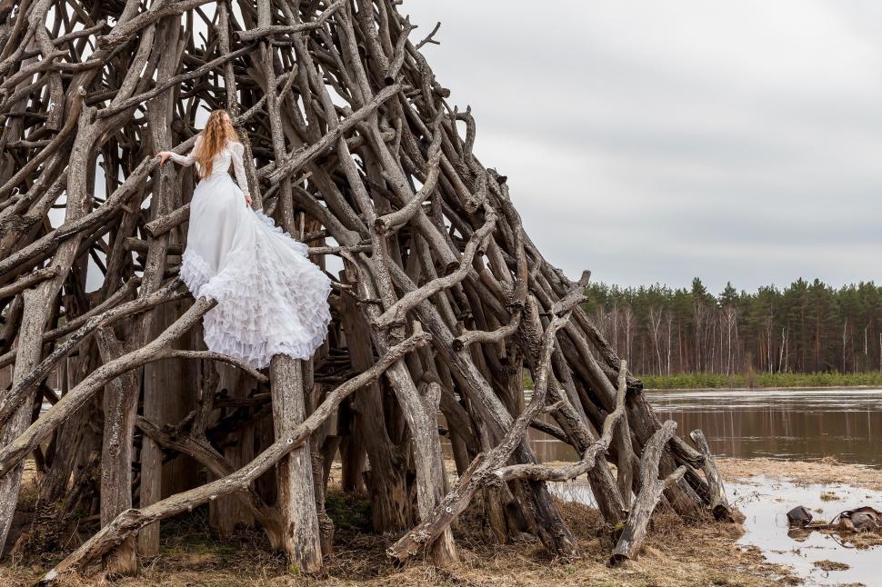 Free Image of Woman in white wedding dress climbing a large wooden structure outdoors 