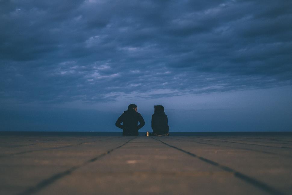 Free Image of Silhouetted couple seated on boardwalk under dark, cloudy evening sky. 
