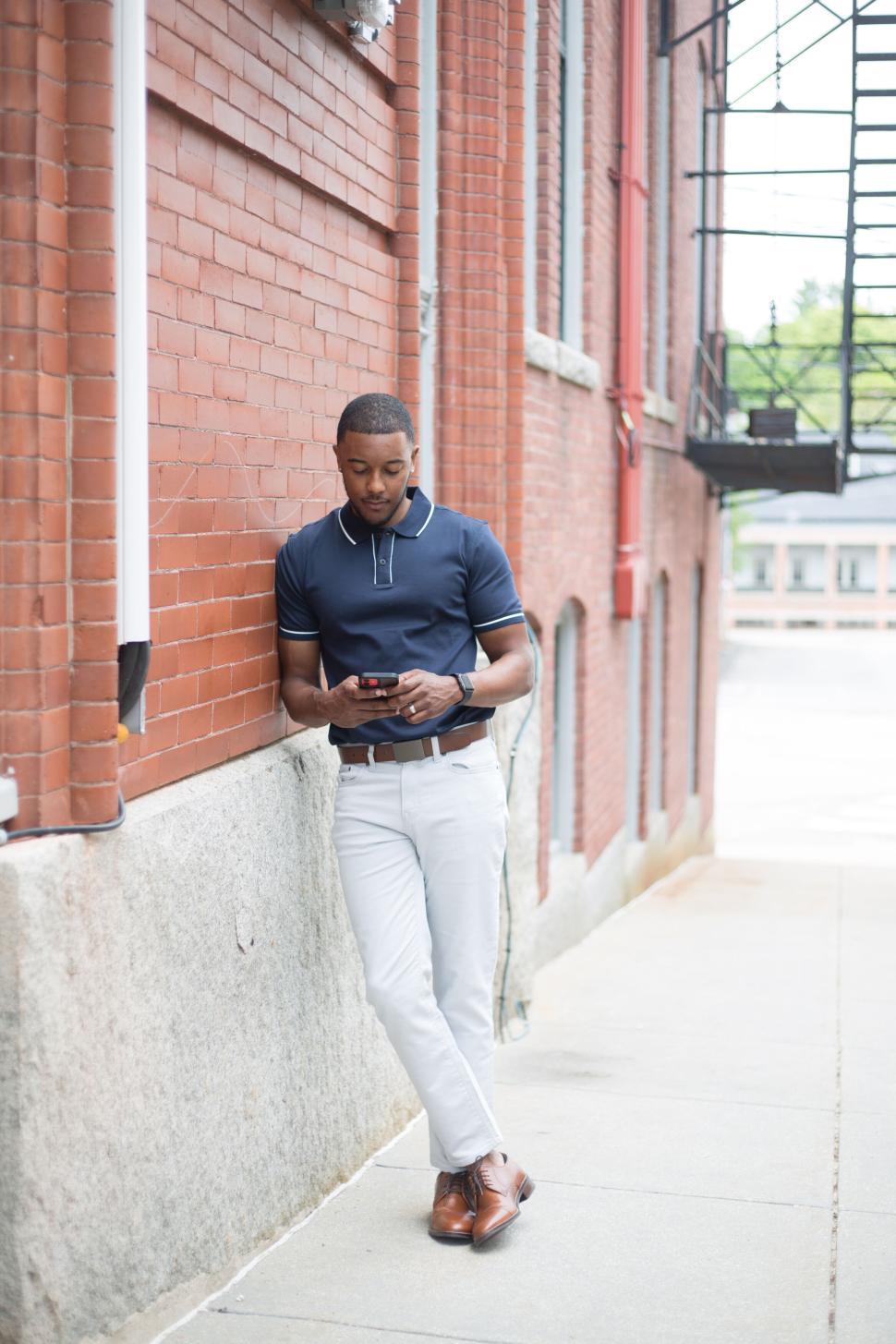Free Image of Casually dressed man leaning against brick wall using smartphone 