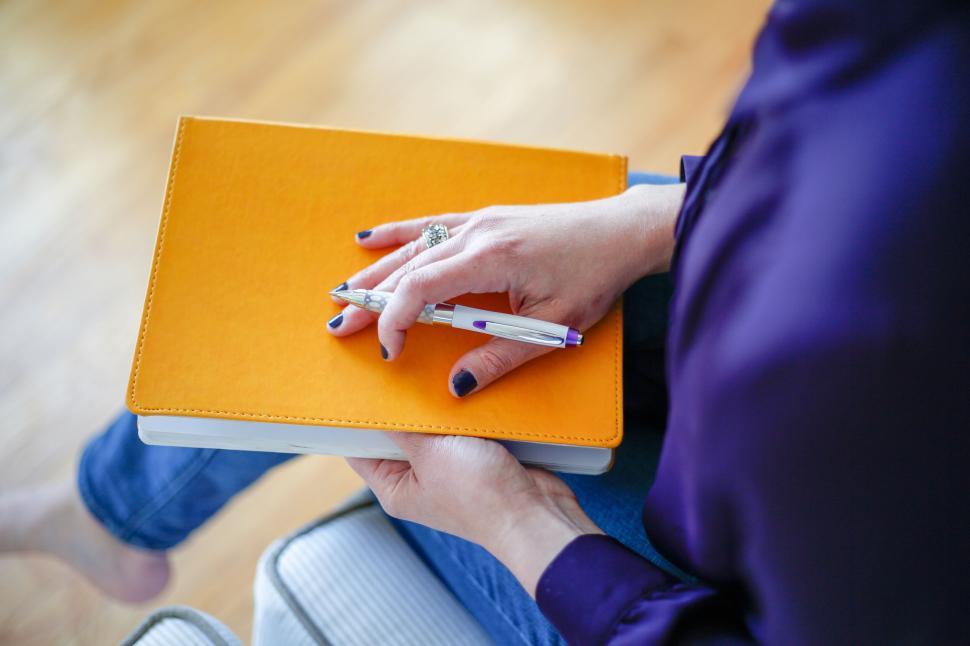 Free Image of Person Holding Orange Book and Pen, Seated on Wooden Surface 