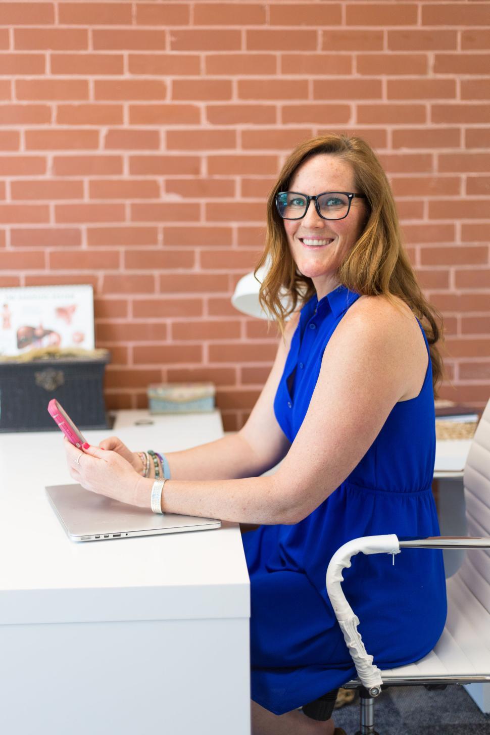 Free Image of Red-haired woman in blue dress smiling,holding phone in office 