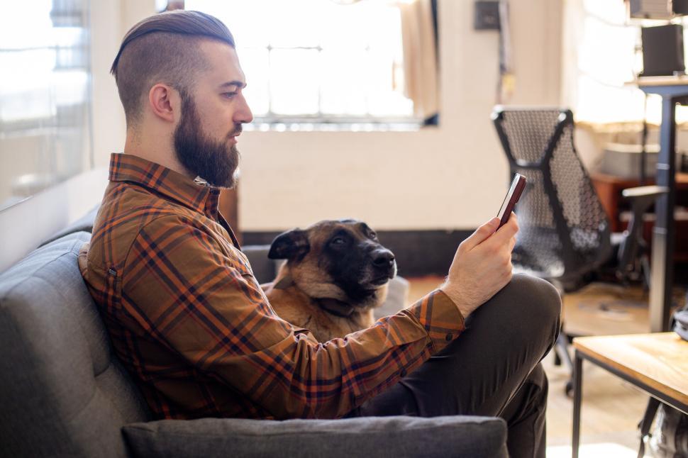 Free Image of Man with dog seated on couch using smartphone in bright room 