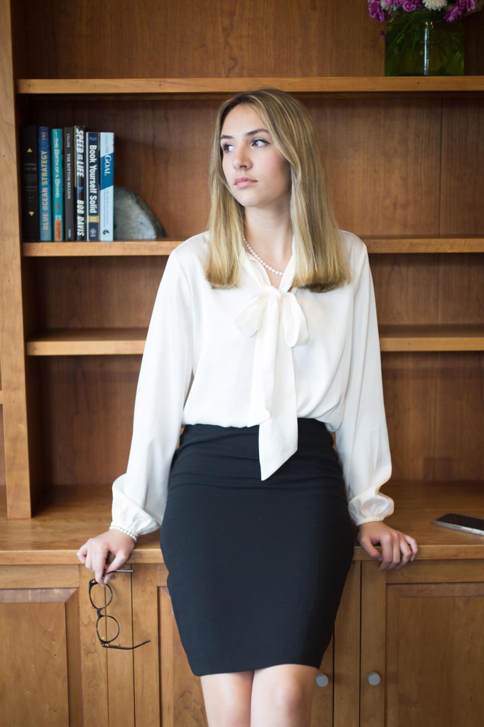 Free Image of Woman standing against bookshelf wearing white blouse and skirt. 