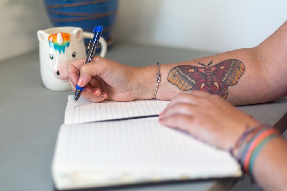 Free Image of Person writing in notebook with tattooed arm and unicorn mug 
