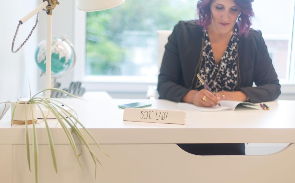 Free Image of Woman writing at a white desk with a  Boss Lady  sign and lamp. 