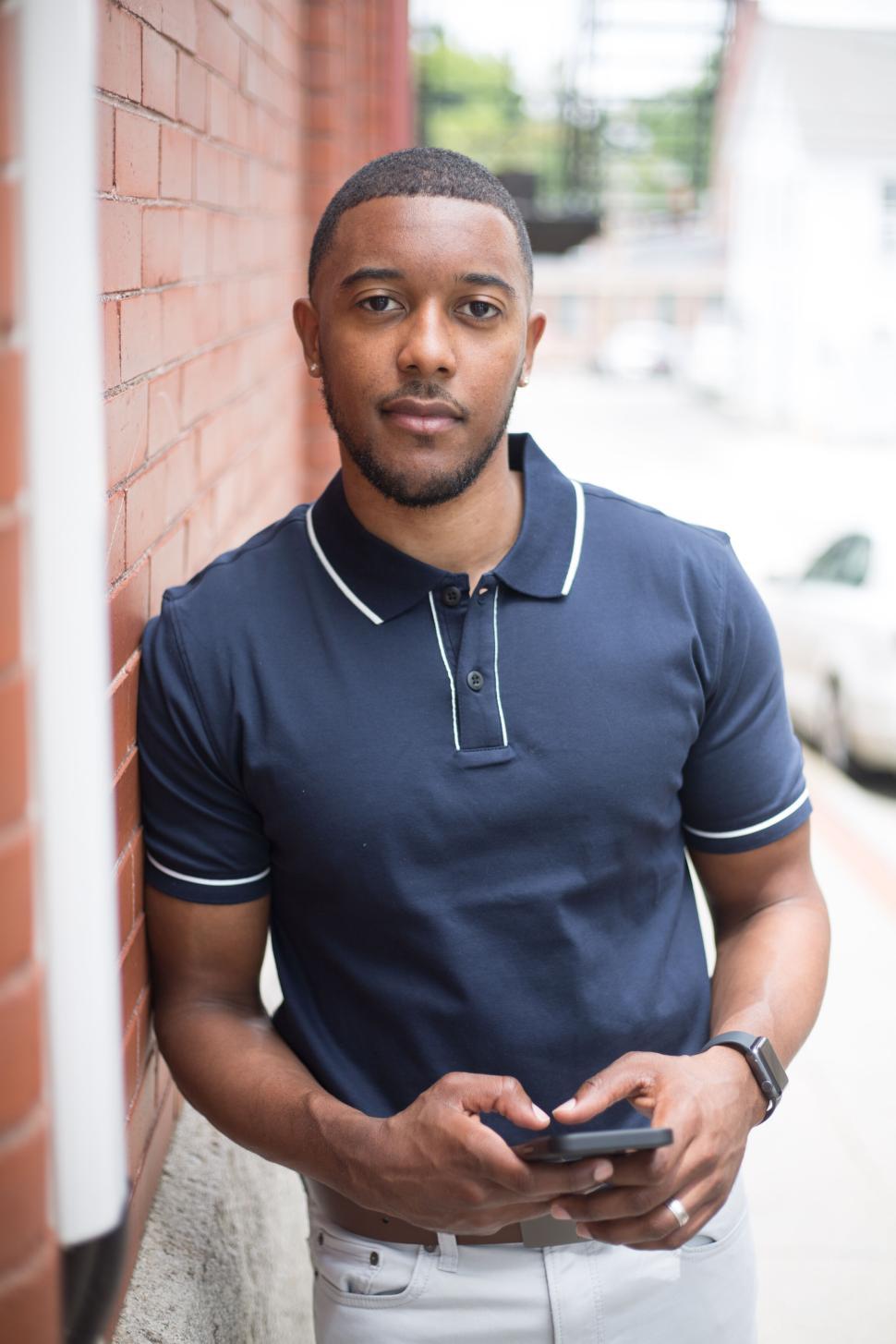 Free Image of Man in casual attire leaning on brick wall, holding smartphone. 
