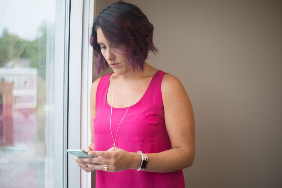 Free Image of Woman in pink top texting on her smartphone by a window indoors. 