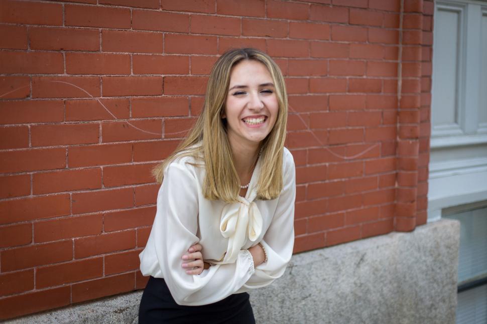 Free Image of Smiling woman in white blouse poses against a brick wall 