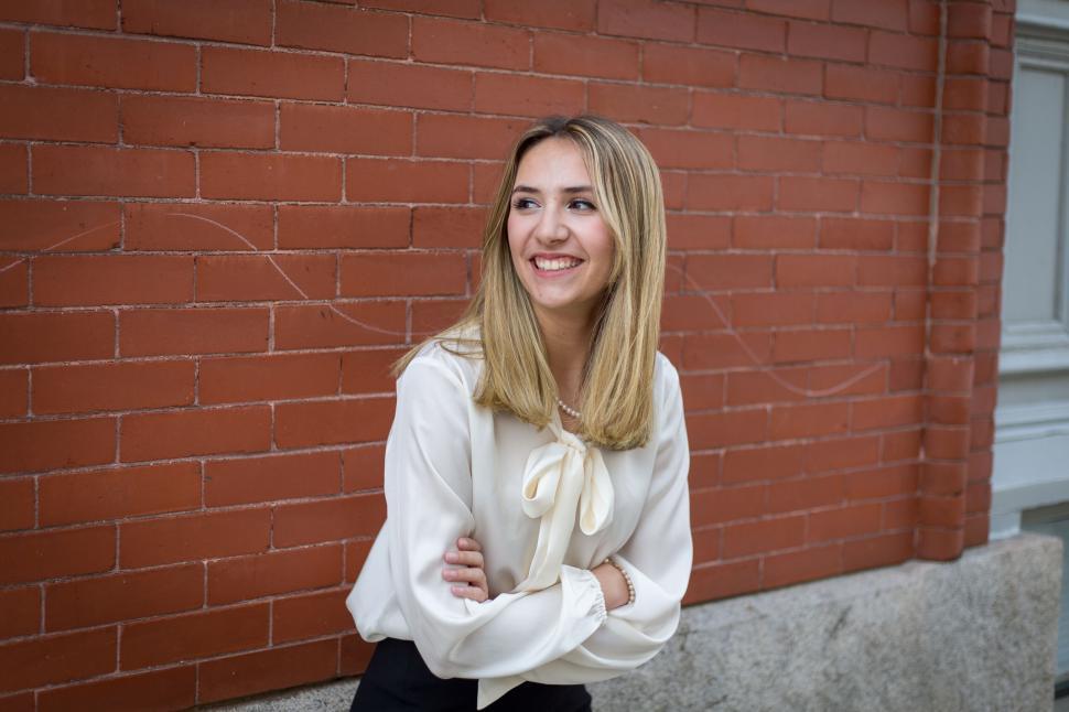 Free Image of Smiling woman in a white blouse poses against a brick wall 