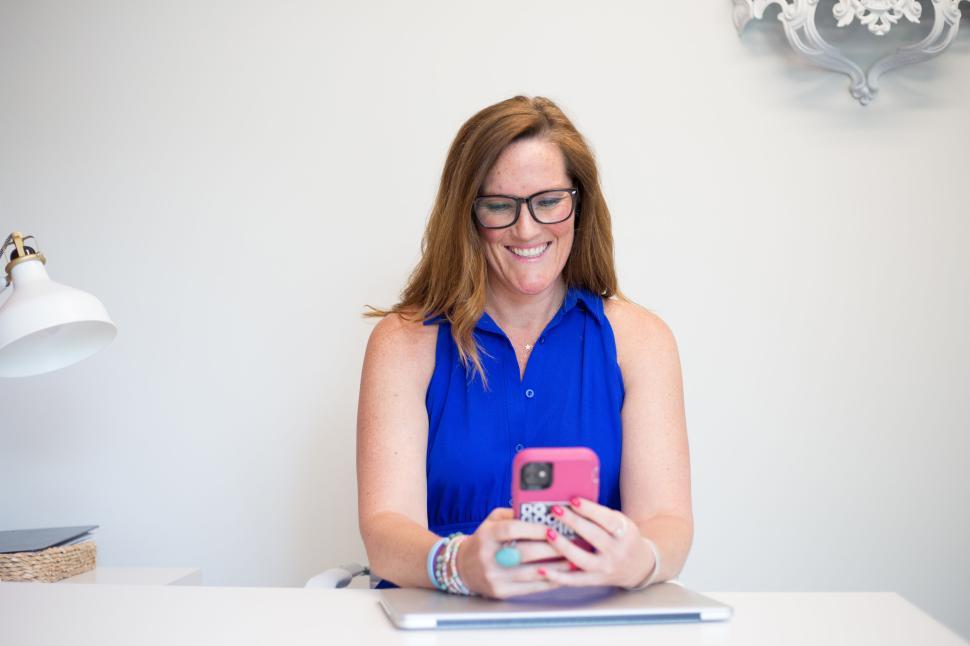 Free Image of Smiling woman in blue dress using smartphone at desk 