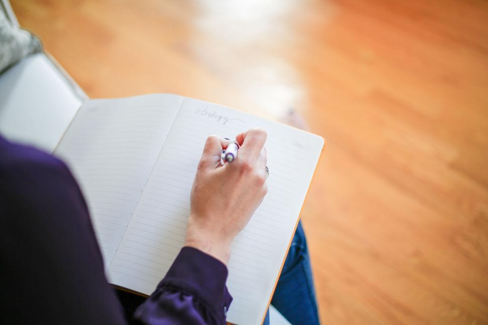 Free Image of Person writing in a notebook with pen on a wooden floor background. 